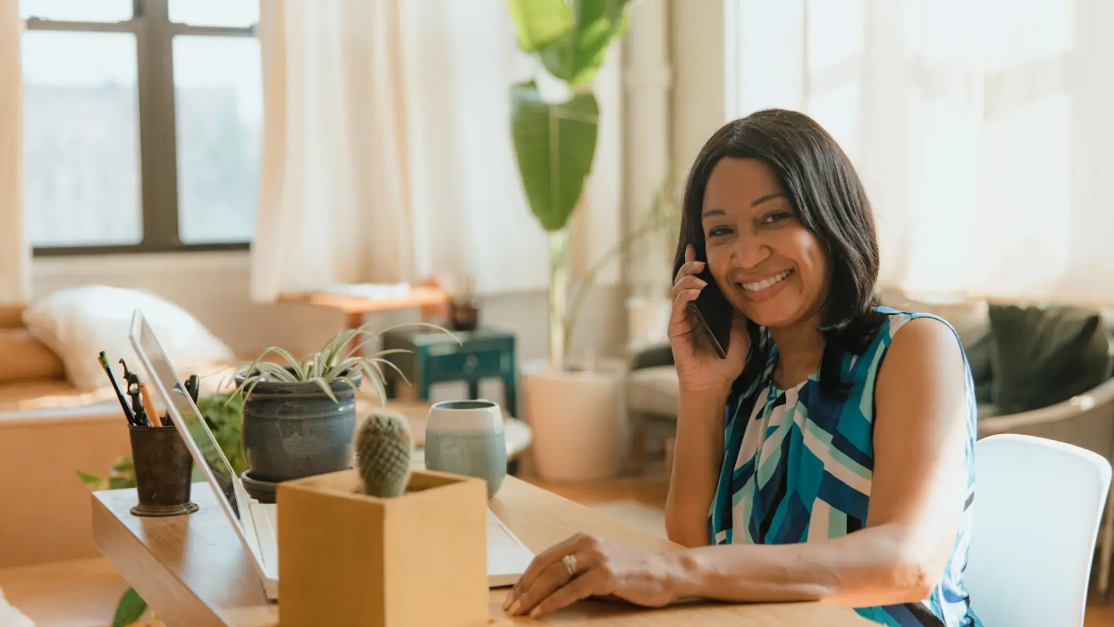 Mulher sorri ao falar ao celular sentada numa mesa de madeira com notebook e vasos de plantas