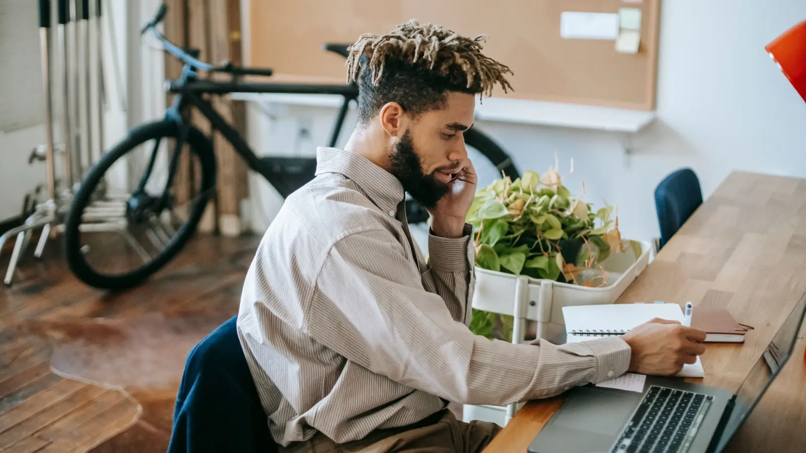 Homem fala ao celular enquanto faz anotações num caderno ao lado do notebook, numa mesa de madeira com plantas