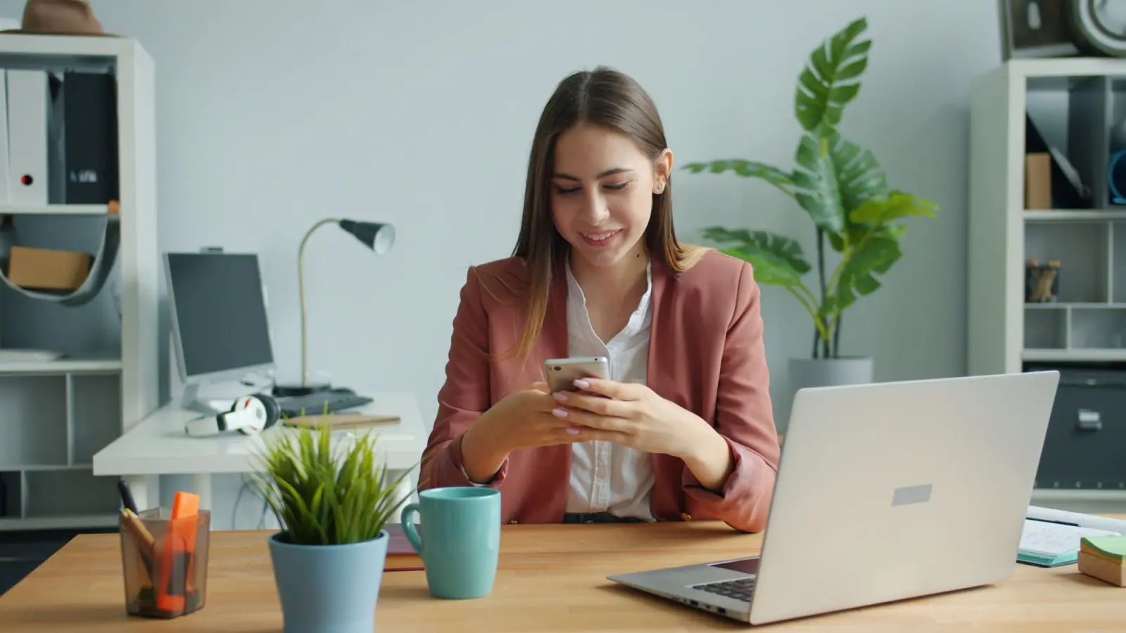Jovem profissional de blazer rosé sorri enquanto usa o celular numa mesa de escritório com notebook, caneca e plantas