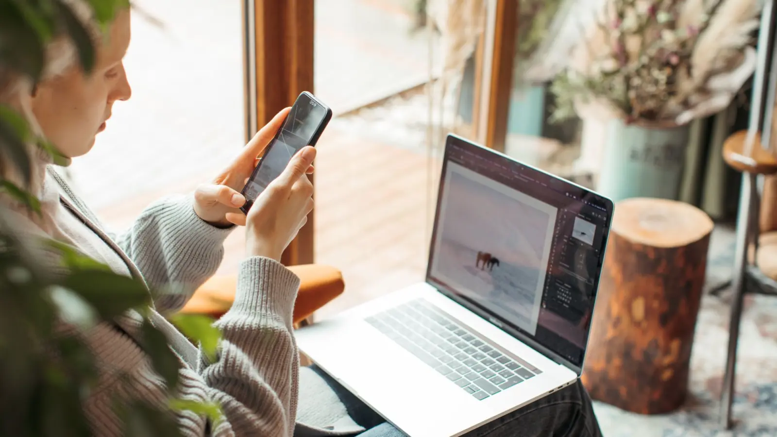 Mulher de suéter usa o celular com um notebook aberto no colo, num ambiente de luz natural e plantas