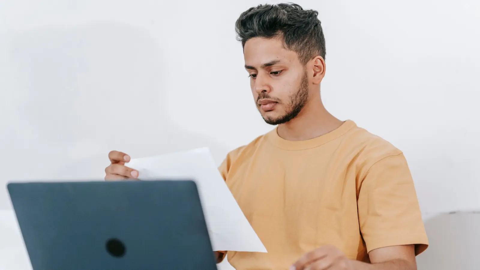 Homem de camiseta amarela confere documentos impressos sentado diante de um notebook
