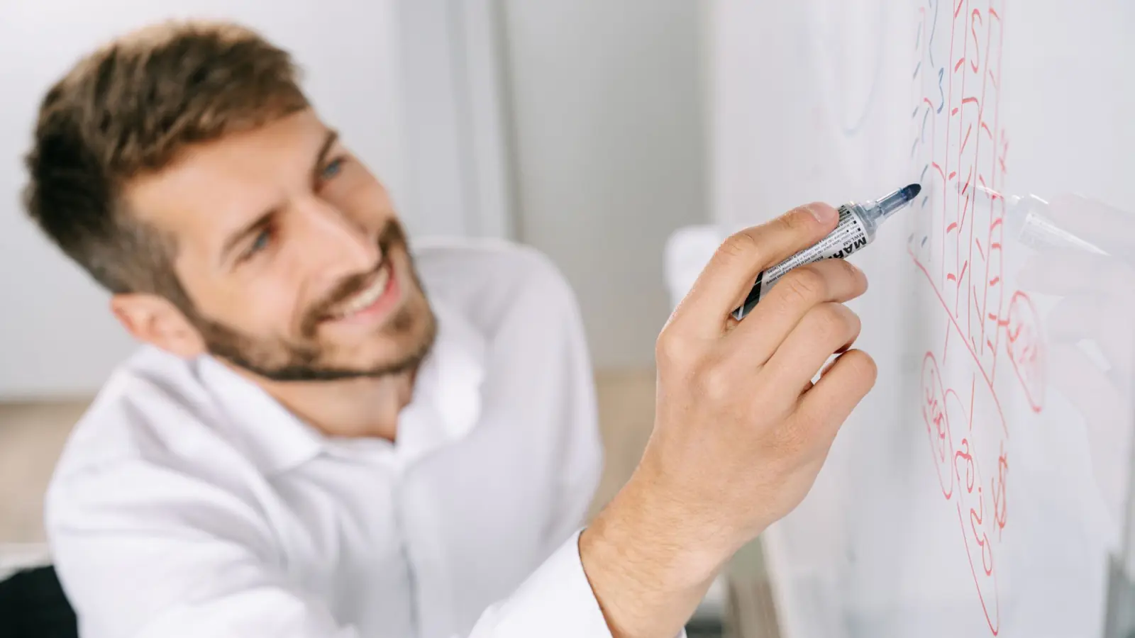 Homem sorridente de camisa branca desenha um fluxo num quadro branco com caneta