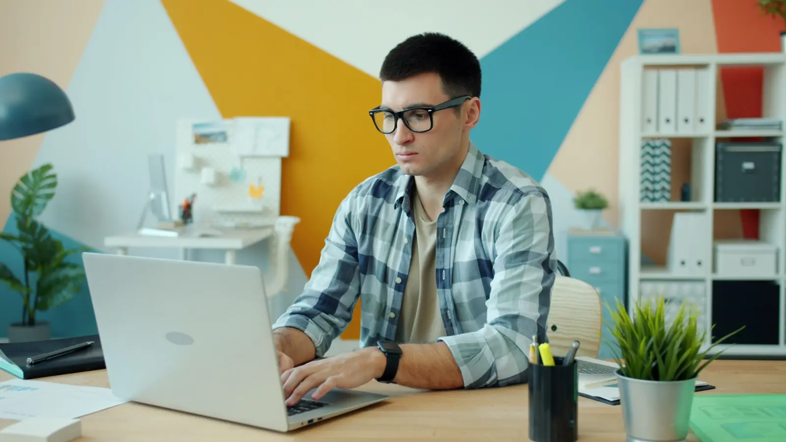 Jovem de óculos e camisa xadrez digita num notebook em um escritório de parede colorida